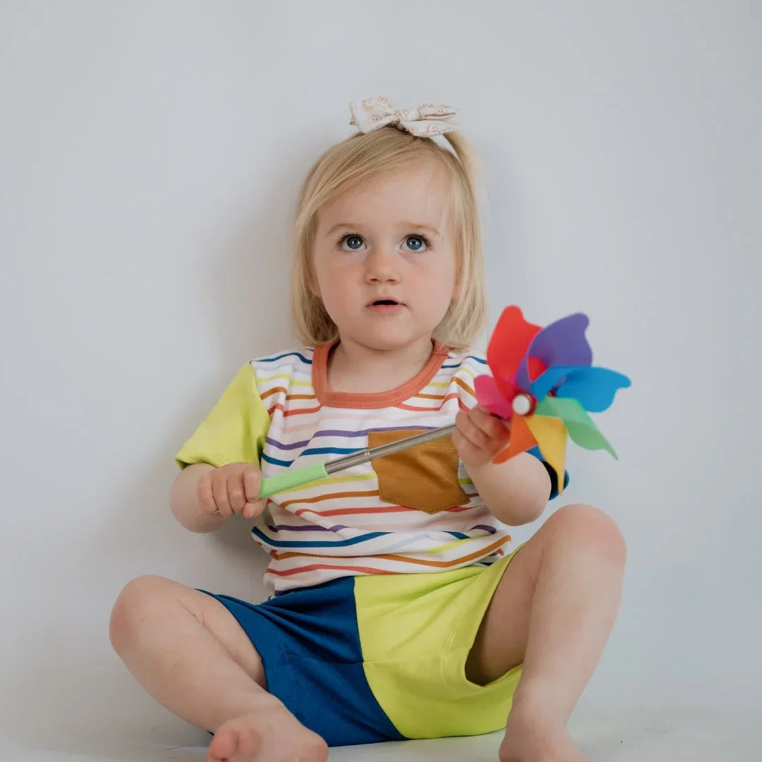 Toddler in striped shirt and shorts holding rainbow pinwheel against white background