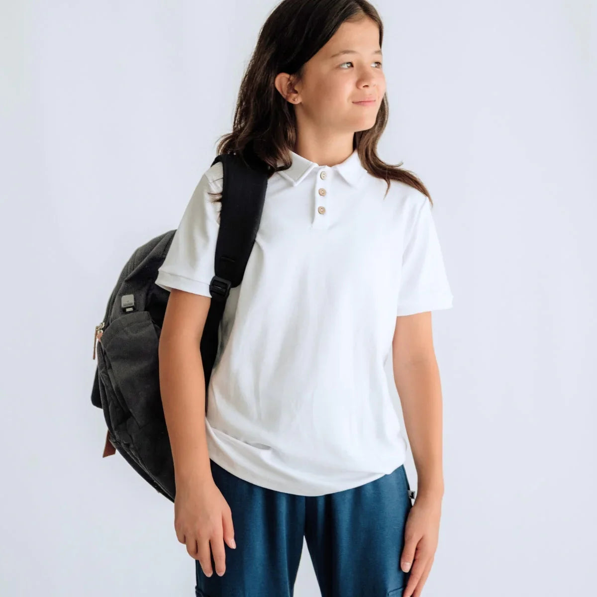 Smiling student in white polo shirt, blue pants, and black backpack on white background