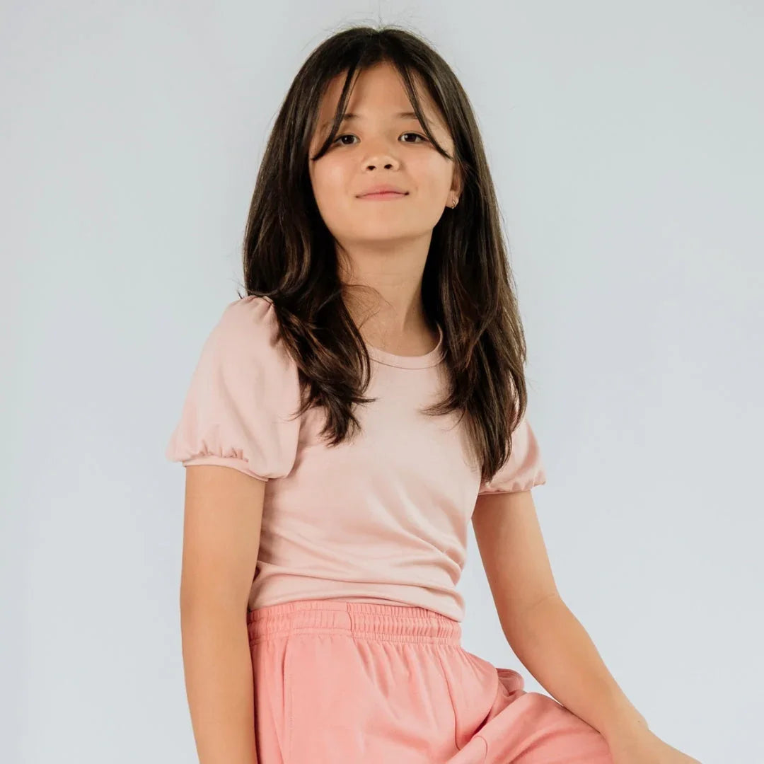Girl wearing light pink puffed shoulder t-shirt and pink shorts against plain background