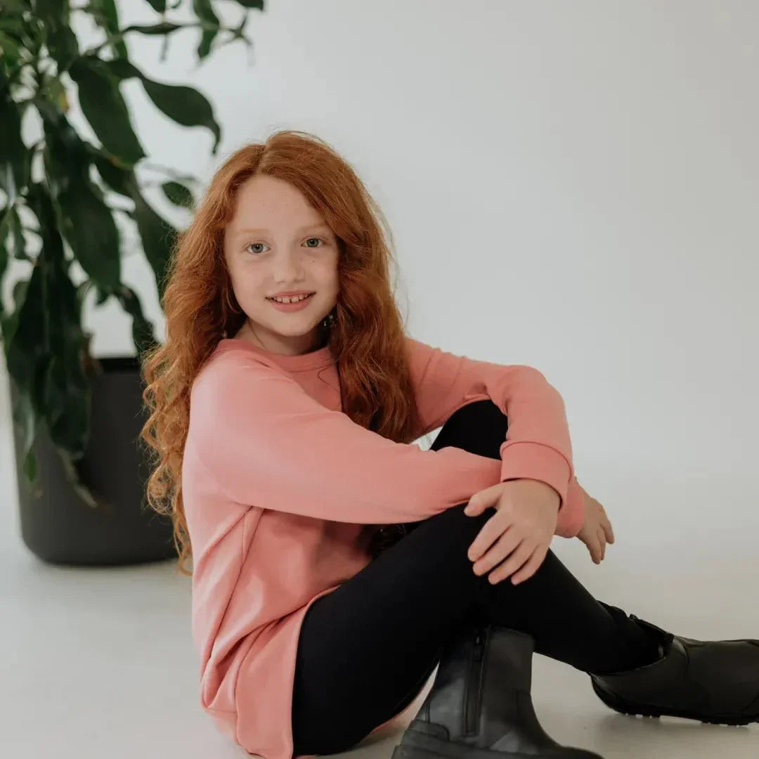 Girl in pink bamboo pullover and black leggings sitting indoors, plant in background