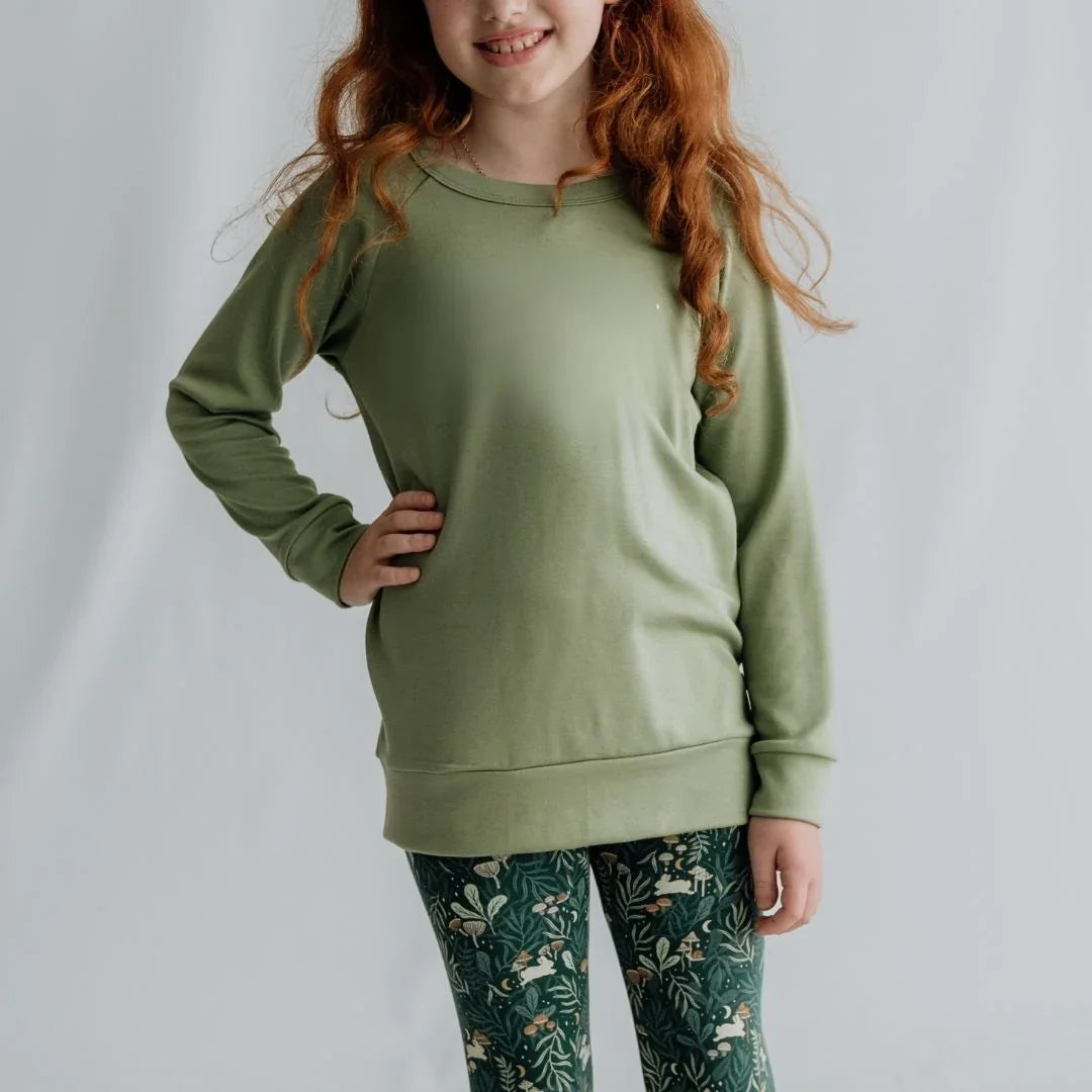 Girl wearing green sweatshirt and patterned leggings standing in front of white background