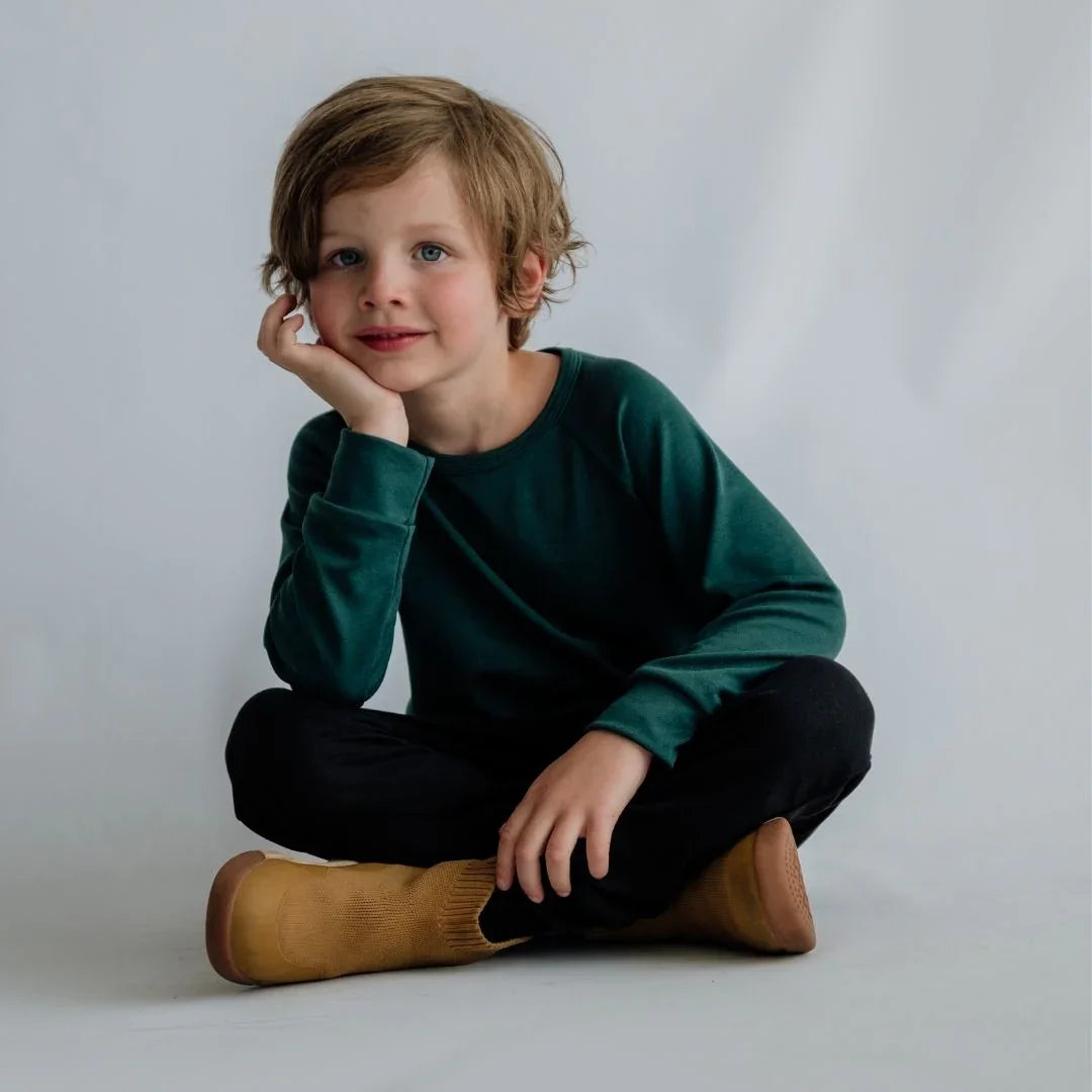 Smiling child in green sweatshirt, black pants, and tan boots sitting on white background