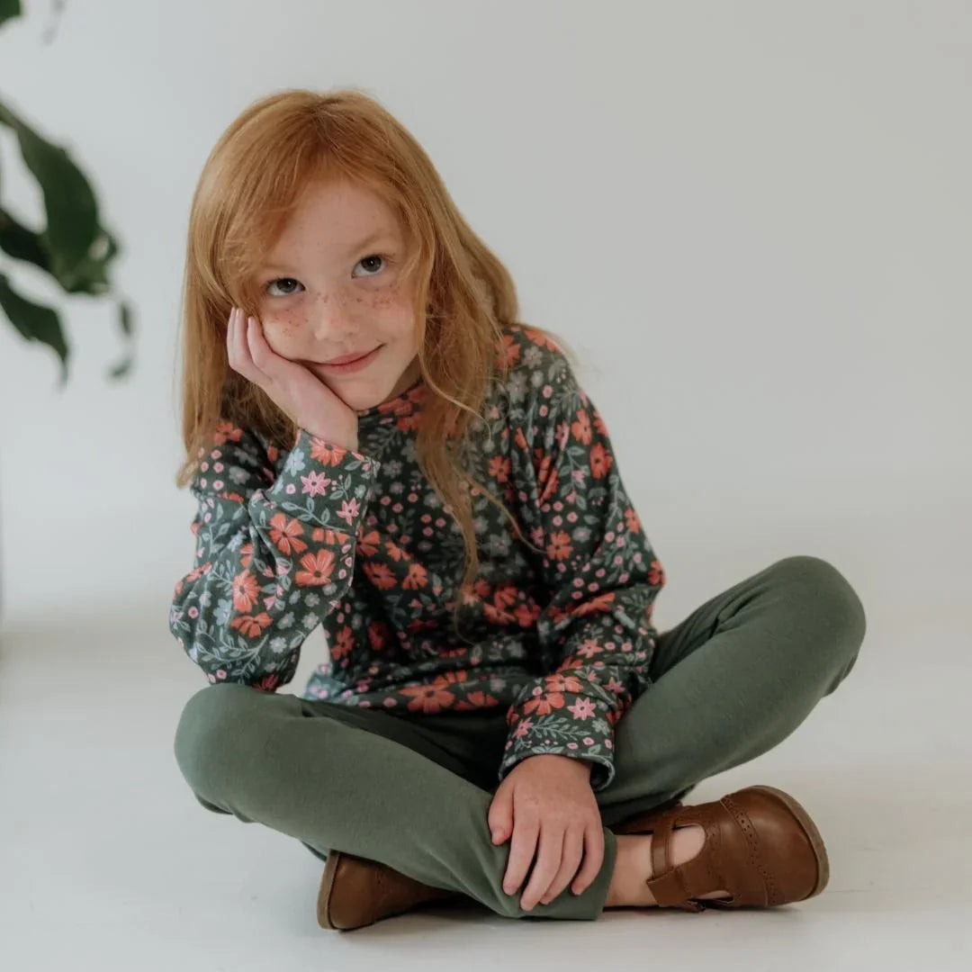 Smiling child in floral bamboo raglan pullover and green pants sitting on floor