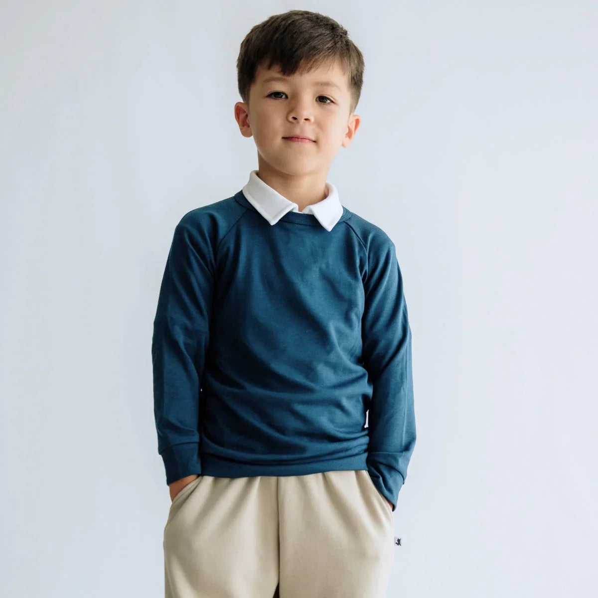 Young boy in blue long sleeve shirt and beige pants standing against a white background.