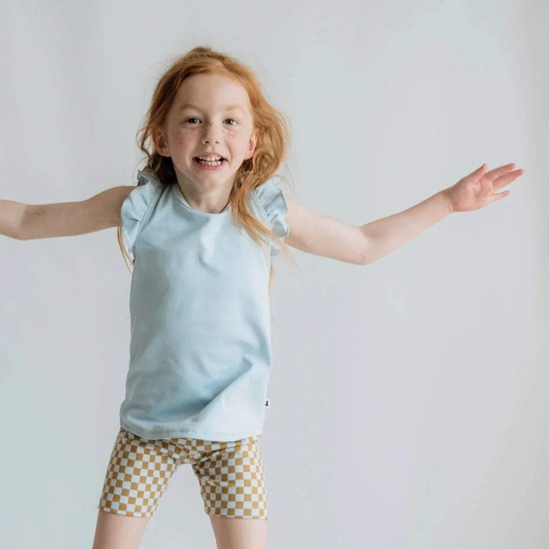 Smiling girl in light blue ruffle sleeve top and checkered shorts against white background
