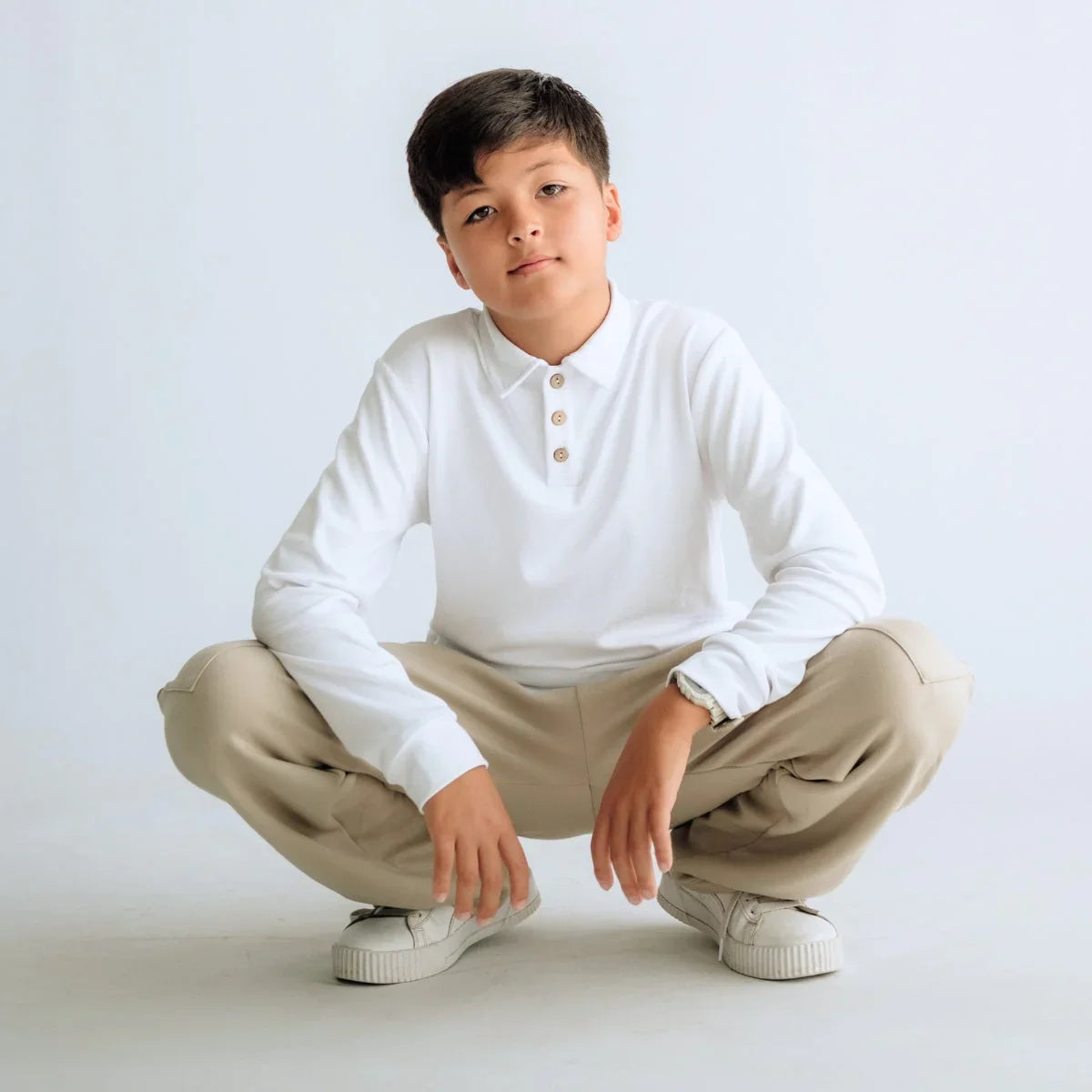 Boy in white long sleeve polo and beige pants squatting in studio, casual fashion look
