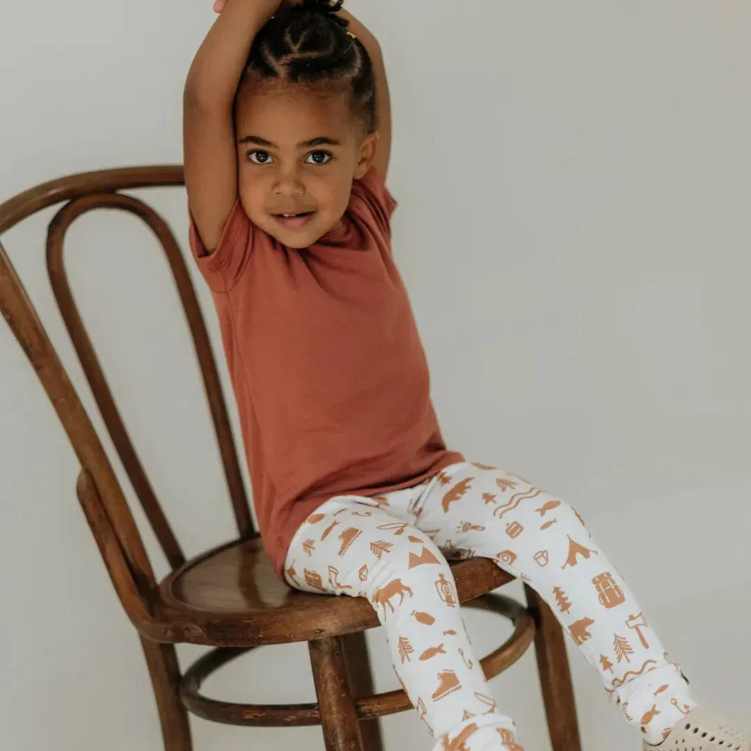 Smiling child in rust bamboo t-shirt and printed leggings sitting on wooden chair indoors