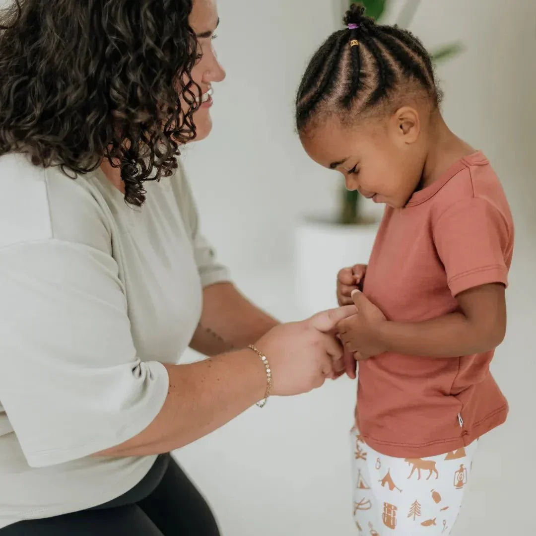 Smiling child in a rust bamboo t-shirt and printed leggings with adult in neutral top indoors