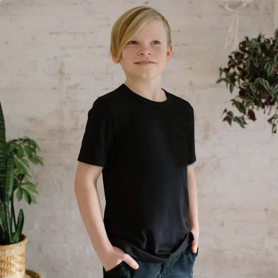 Smiling youth wearing black t-shirt and dark pants, standing indoors with plants nearby