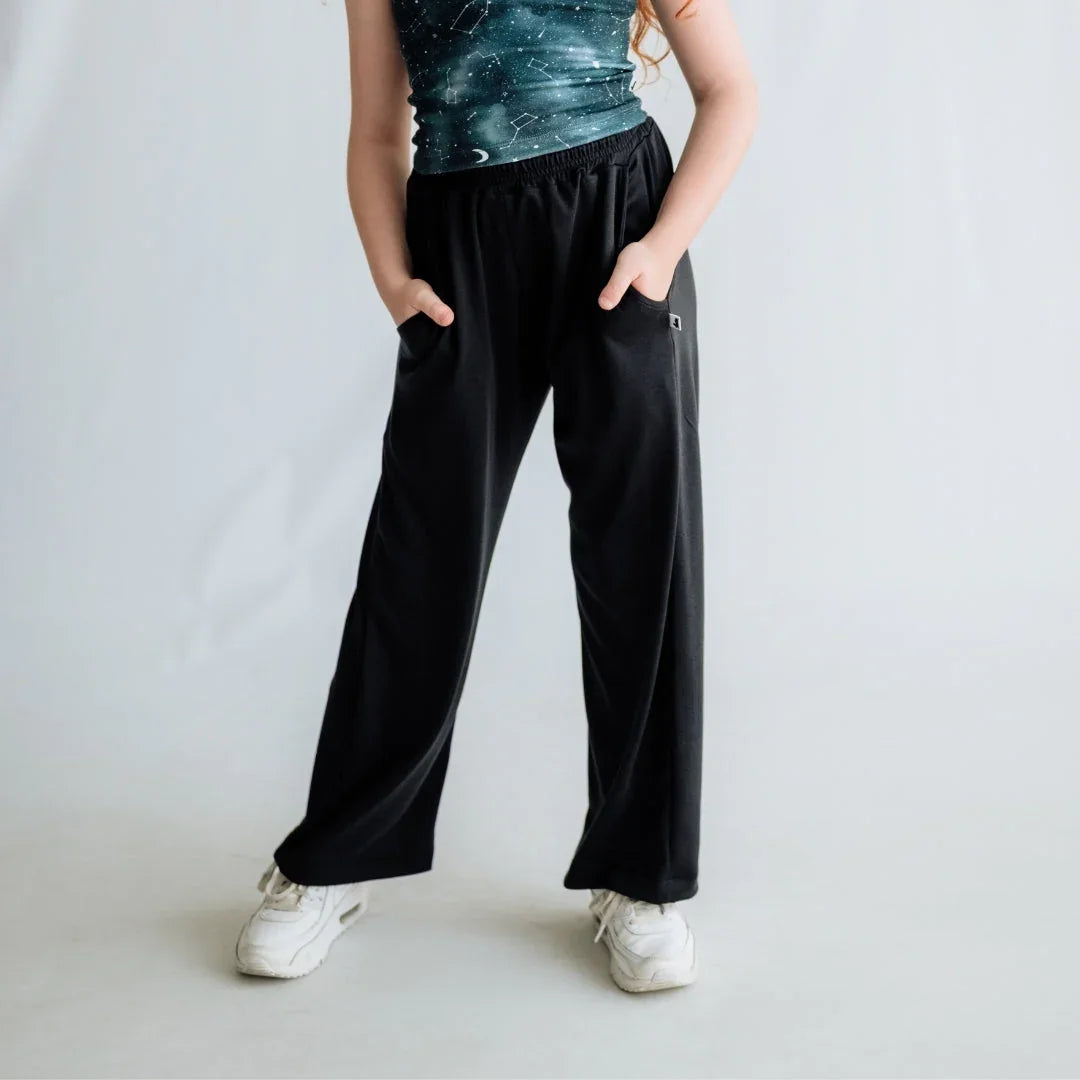 Girl wearing black wide leg pants and white sneakers, hands in pockets, studio background