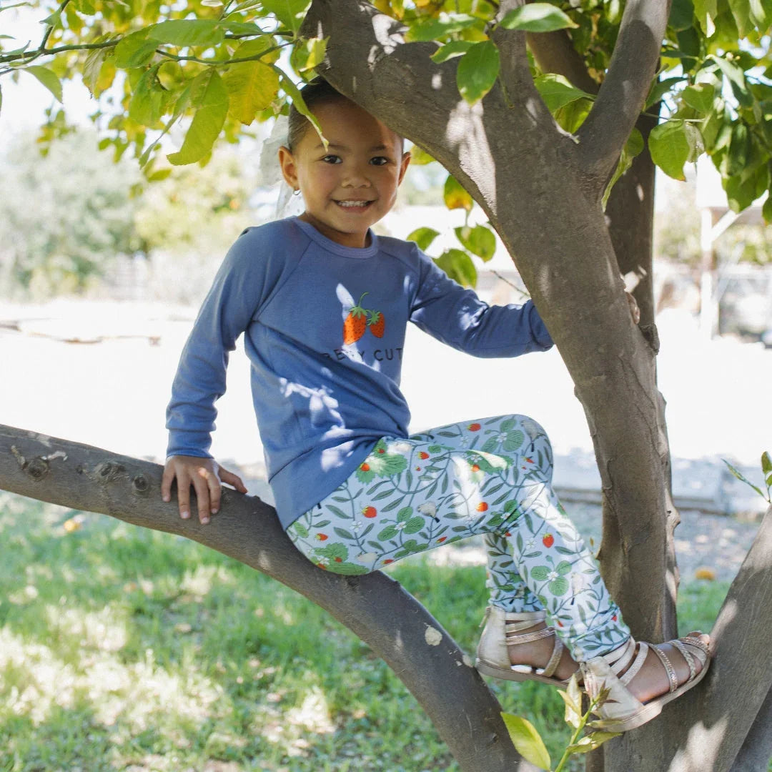 Smiling child in blue top and floral leggings sitting on a tree branch outdoors
