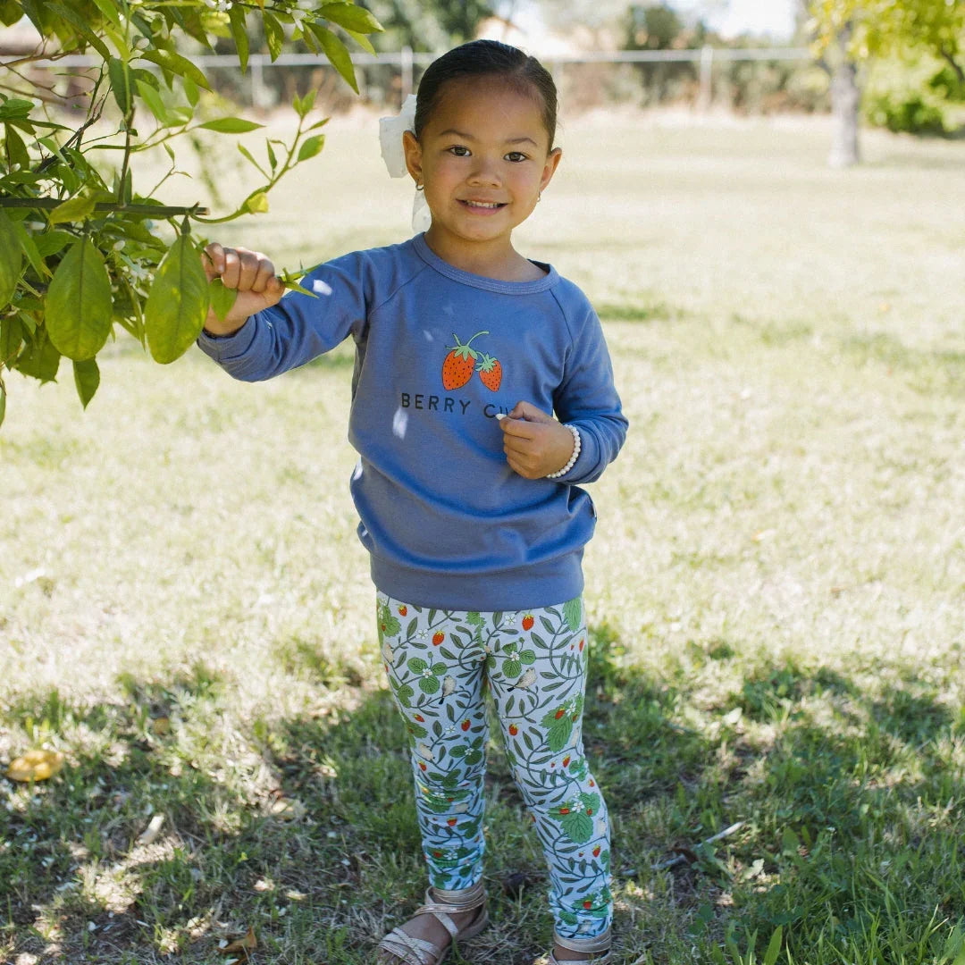 Smiling girl in blue Berry Cute sweatshirt and printed leggings standing by a leafy tree outdoors