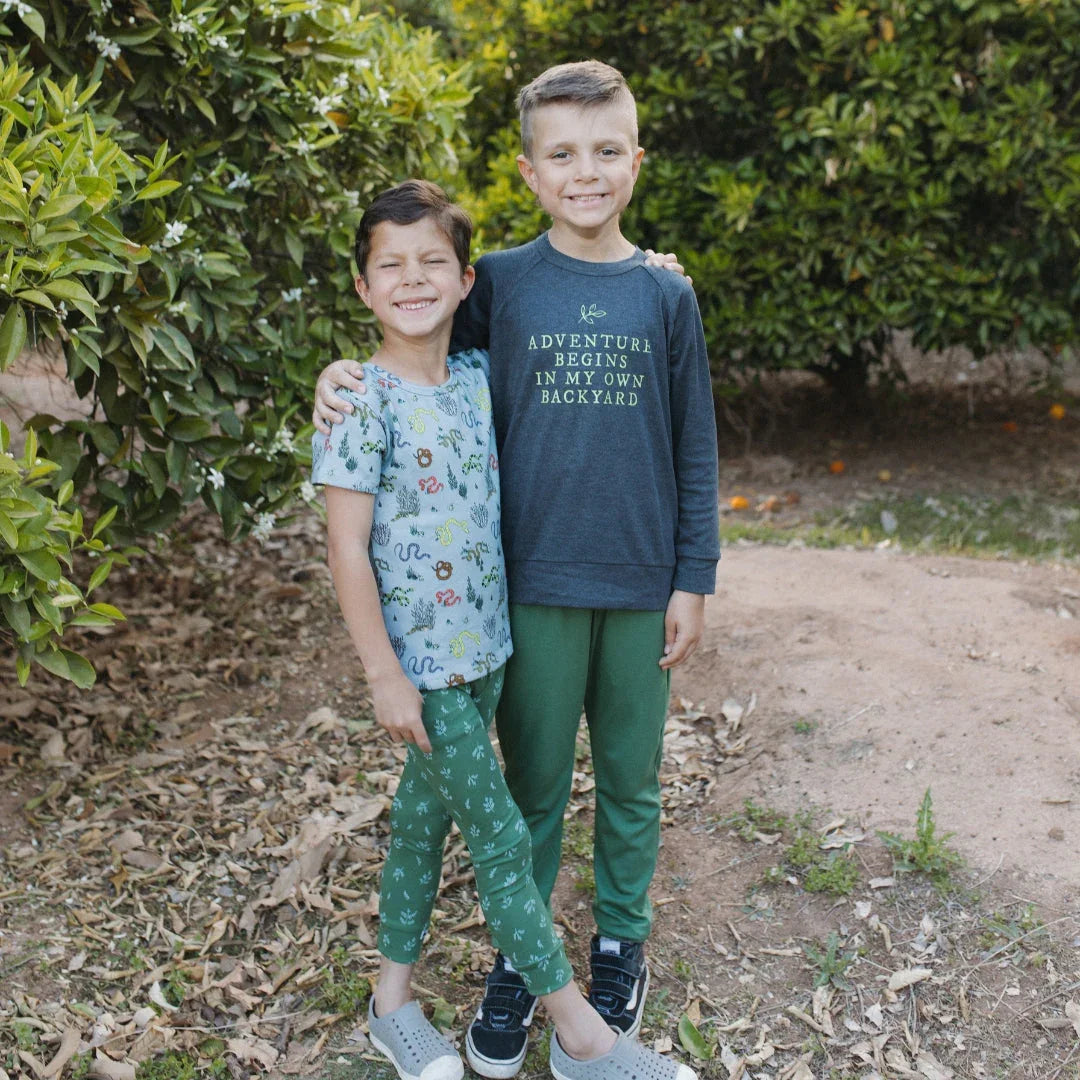 Two smiling boys in casual clothes standing outdoors by green bushes and dirt ground