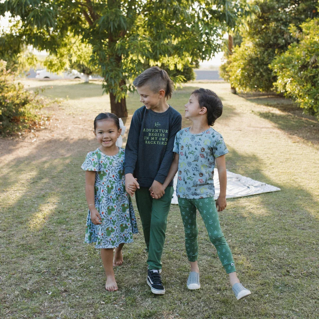 Three smiling children in nature-themed outfits play together outdoors on grass under trees.