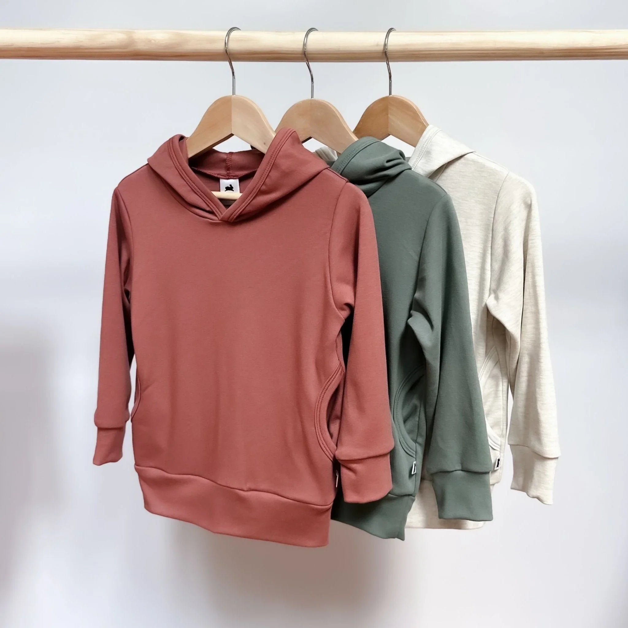 Three youth lightweight bamboo hoodies in pink, green, and beige on wooden hangers.