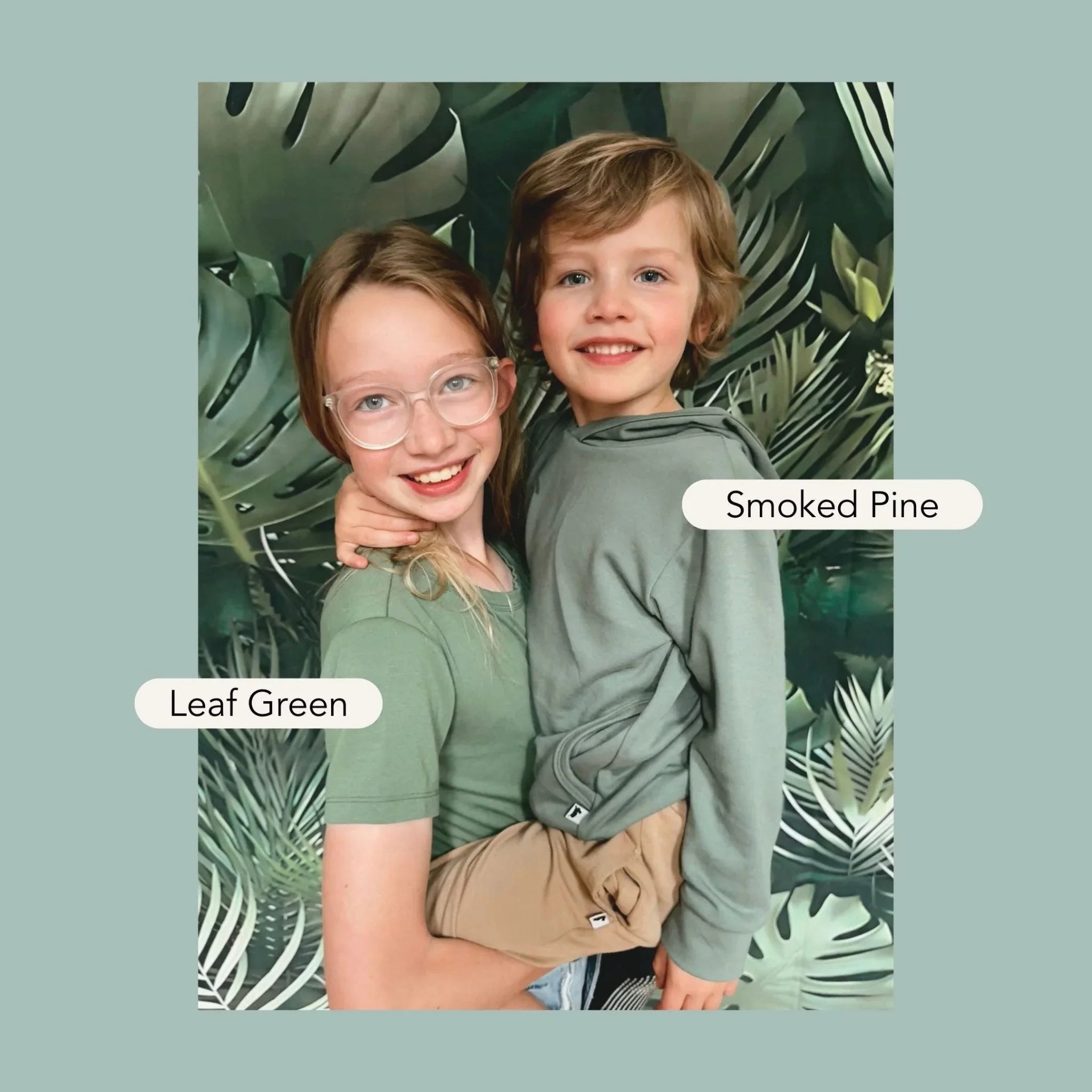 Two kids in green bamboo hoodies, Leaf Green and Smoked Pine, with leafy backdrop