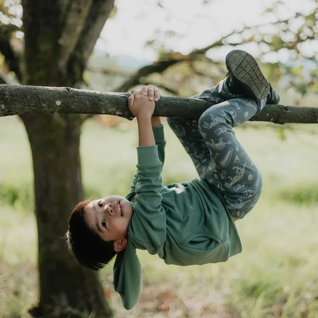Boy in green bamboo hoodie and patterned pants hanging on tree branch outdoors