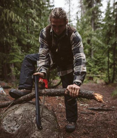 Man using Zippo AxeSaw to cut log outdoors in forest, wearing flannel and vest