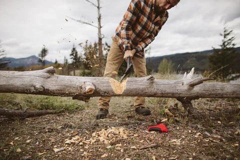 Man using Zippo AxeSaw to cut log outdoors, wearing plaid shirt and khakis, forest background