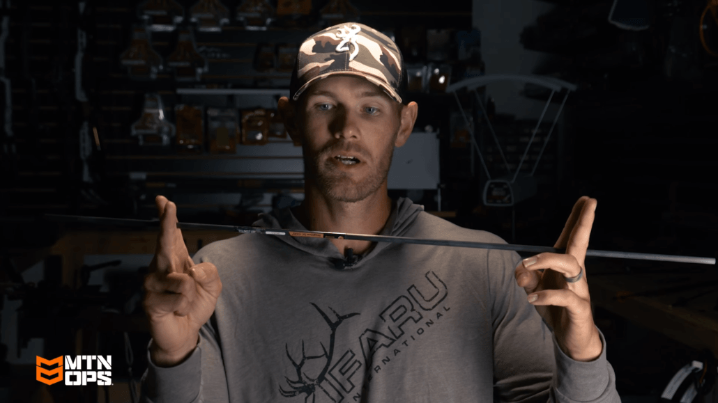 Man in camo hat and Kuiu shirt holding carbon arrow horizontally, archery gear background