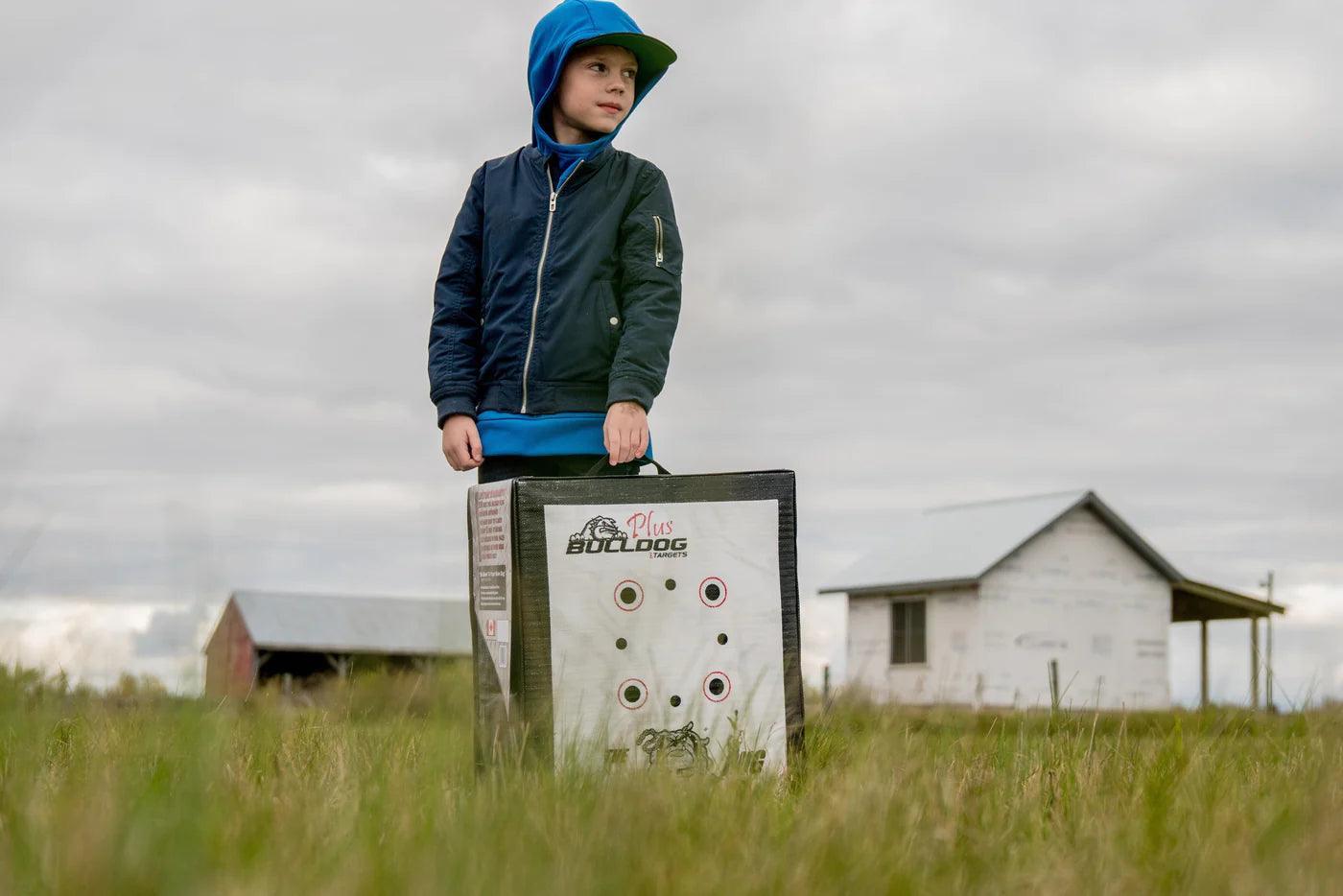Child in blue jacket holding Bulldog archery target outdoors on grassy field with farm buildings