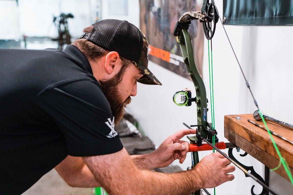 Man tuning a compound bow on workbench with archery equipment indoors