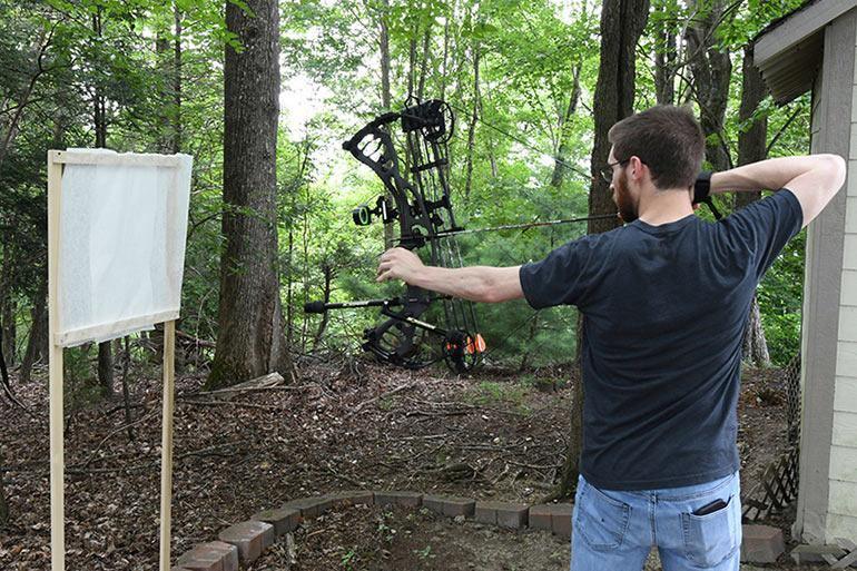 Man paper tuning compound bow outdoors near a white target stand in wooded area