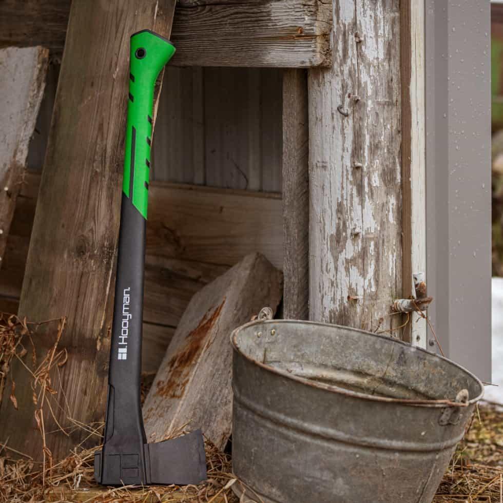 Hooyman chopping axe with green handle leaning on rustic wood near a metal bucket outdoors
