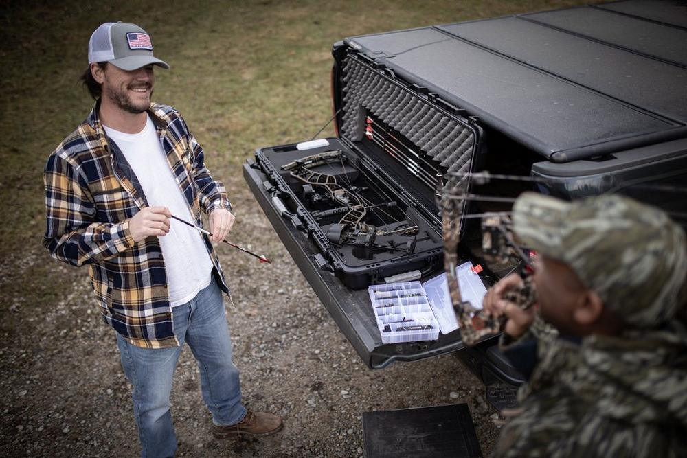Two men with archery gear at a truck with a Plano bow case and arrows outdoors