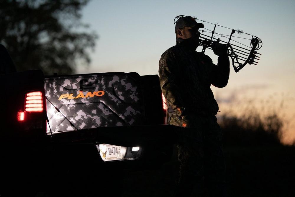 Hunter with compound bow beside truck and Plano camo archery bow case at sunset outdoors