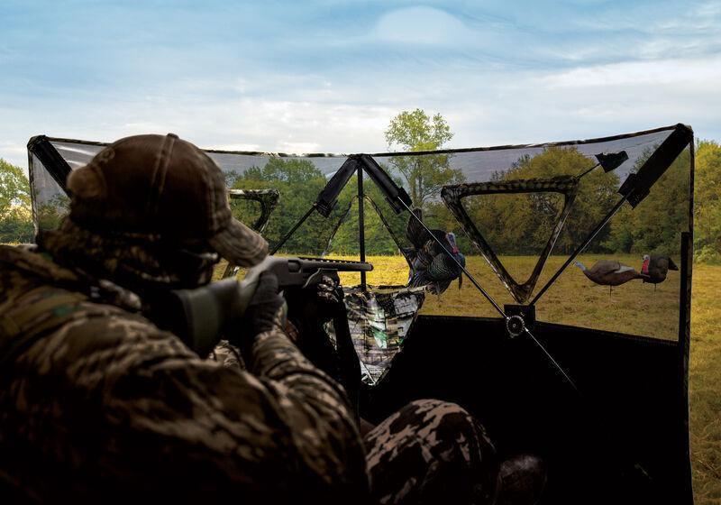 Hunter in camouflage aiming from a Primos Double Bull SurroundView stakeout blind in a field