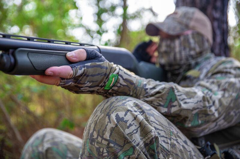 Hunter in camo suit wearing Primos fingerless gloves aiming shotgun in forest