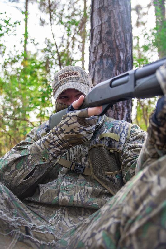 Hunter wearing Primos Stretch Fingerless Gloves in Mossy Oak Bottomland camo aiming shotgun outdoors
