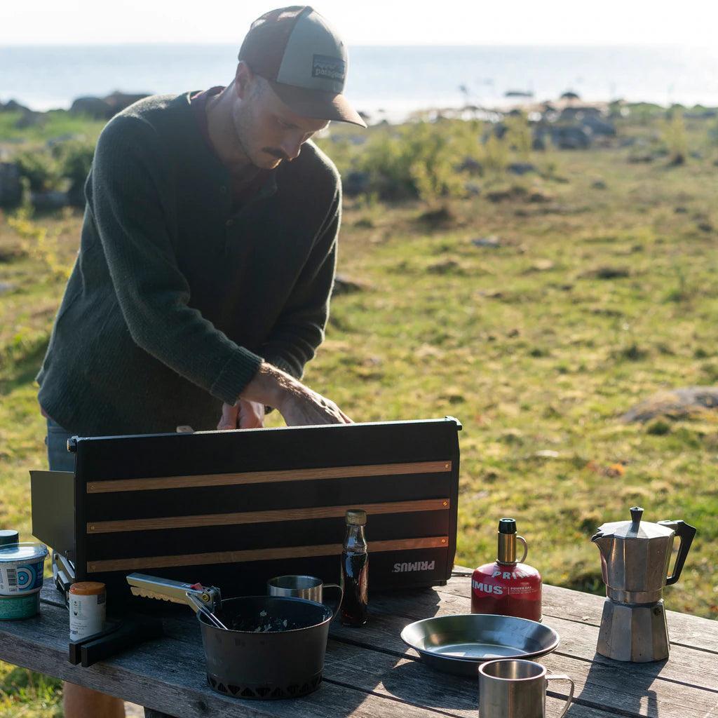 Man using Primus Alika camping stove outdoors on wooden picnic table with kitchenware