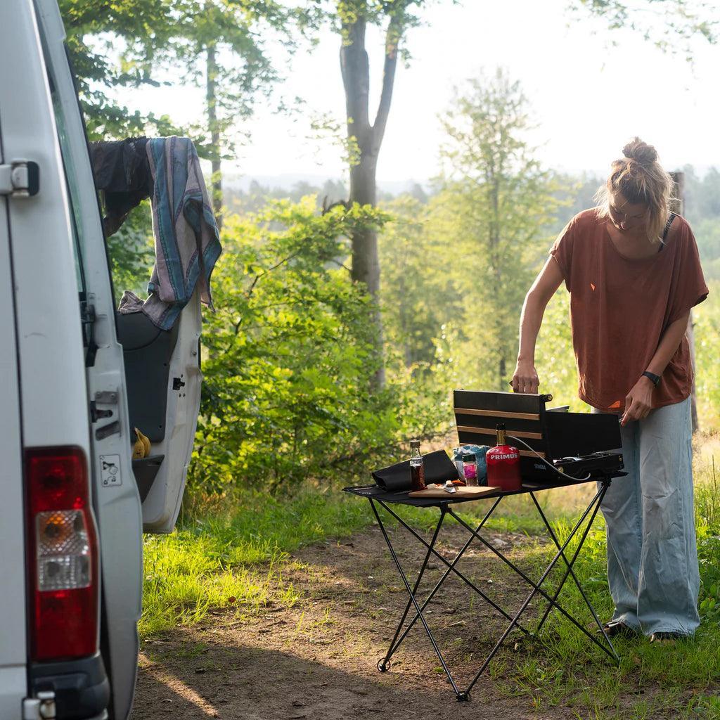 Woman using Primus Alika stove on camping table near van in forest setting