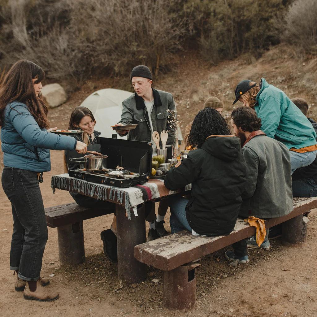 Group camping outdoors cooking on Primus Alika stove at picnic table in nature setting