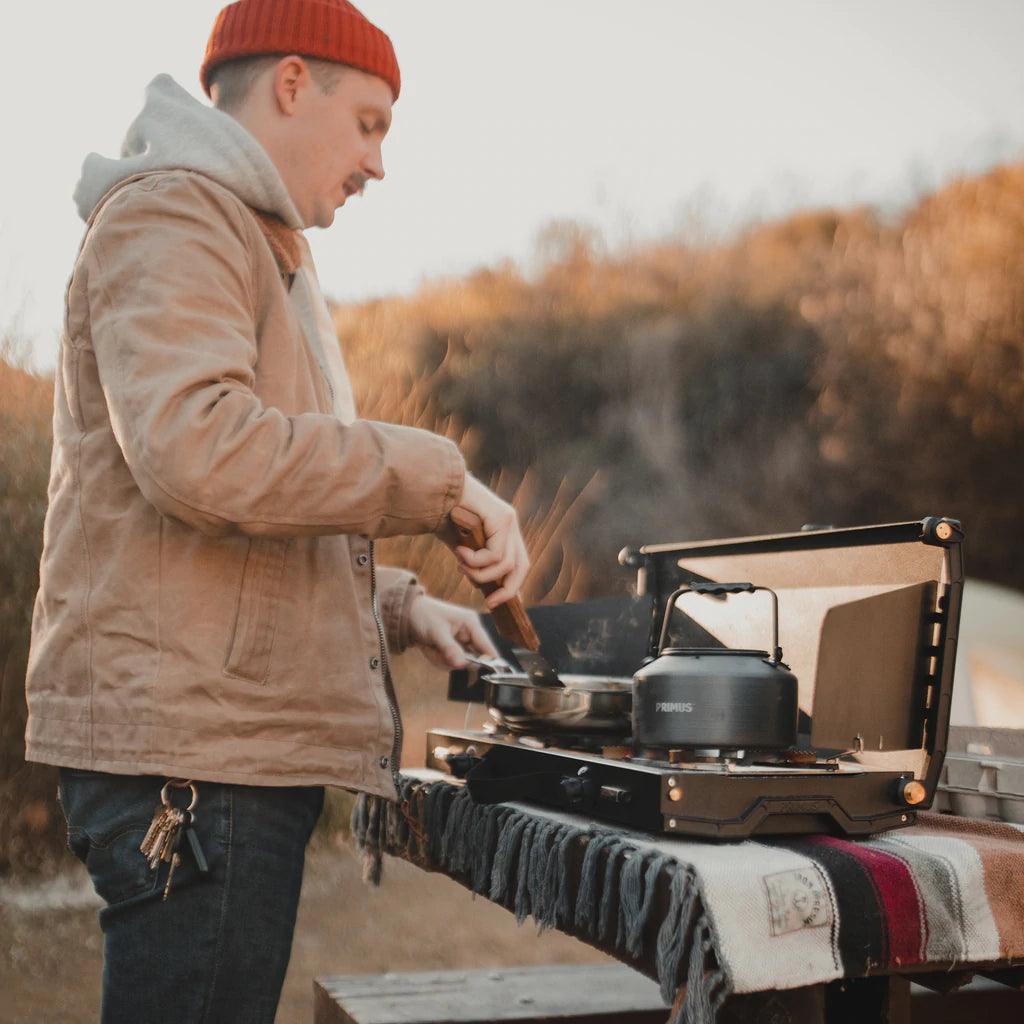 Man cooking outdoors on a Primus Alika camping stove with kettle and skillet