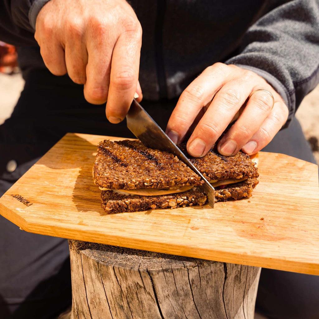 Person slicing sandwich on wooden cutting board outdoors, camping food prep.