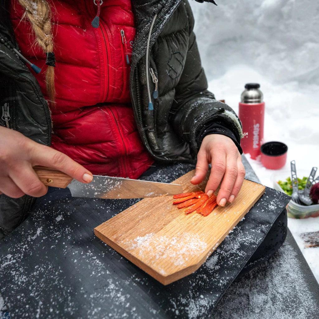 Person slicing red pepper on wooden cutting board outdoors in snowy campsite with Primus thermos