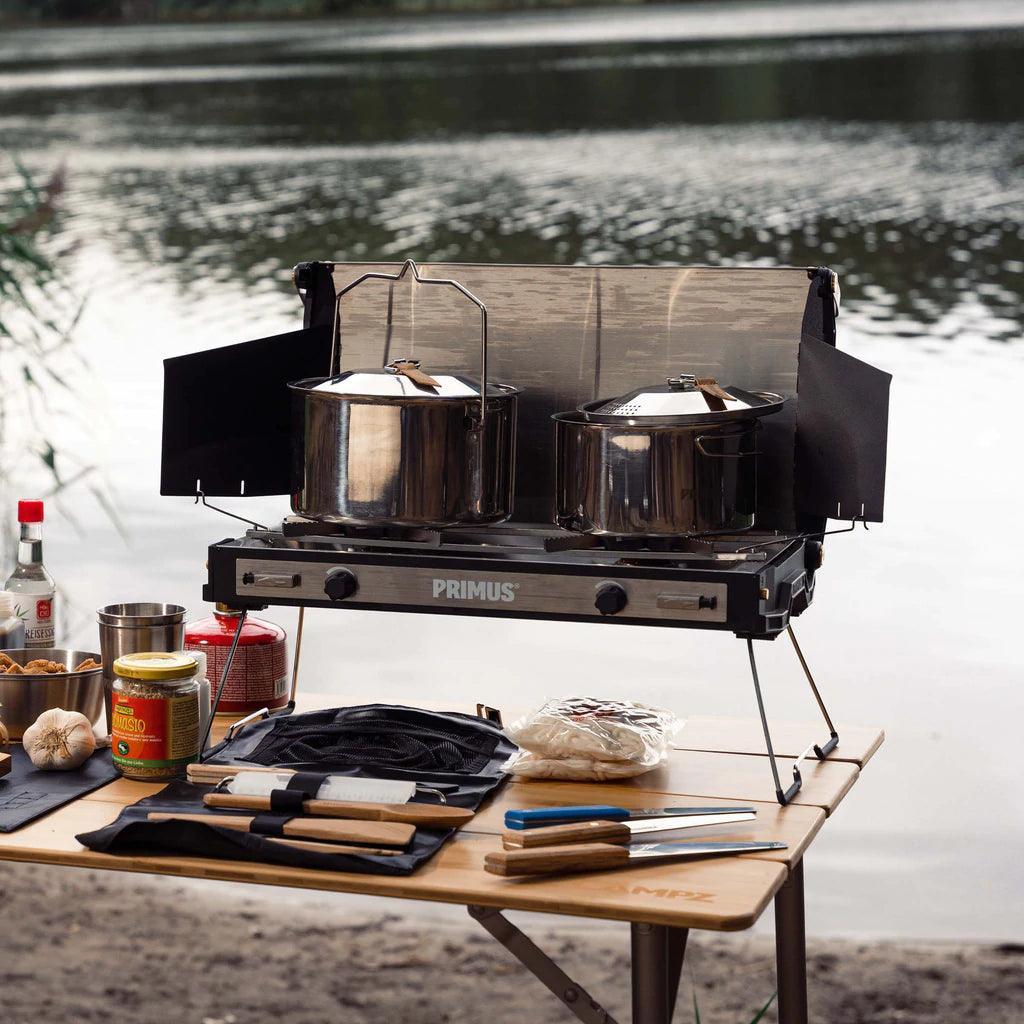 Primus campfire pot set on portable stove with cooking tools by a lakeside outdoor campsite.