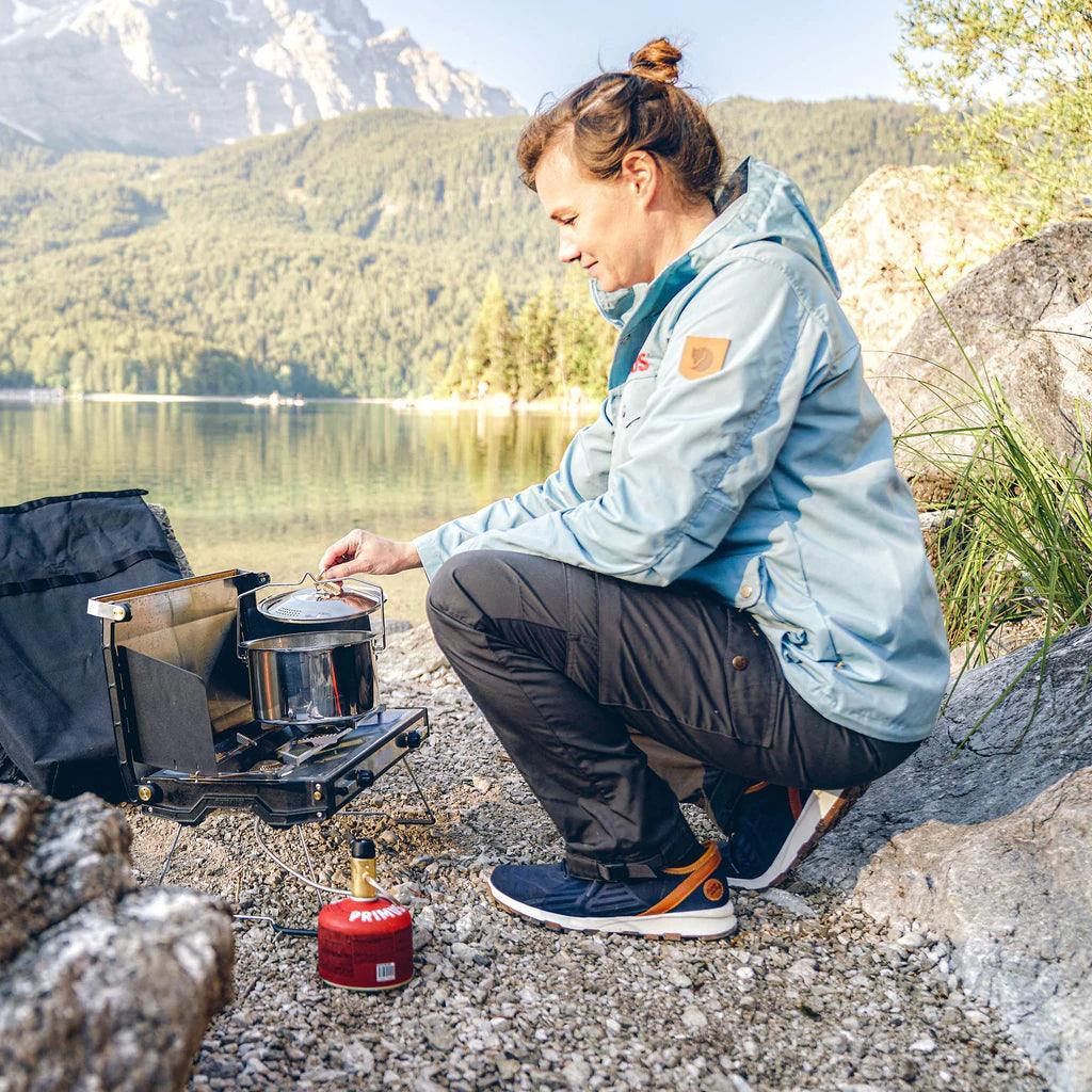 Woman cooking outdoors with Primus Campfire stainless steel pot by a lake in the mountains