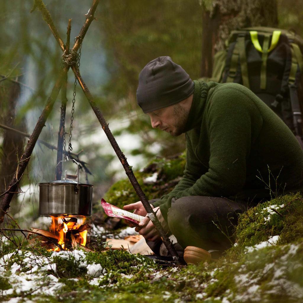 Man with backpack camping in forest, cooking with Primus campfire pot over open fire in snow.