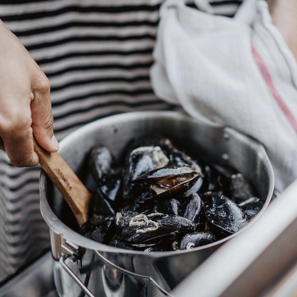Stainless steel Primus campfire pot filled with cooked mussels, hand stirring with wooden spatula.