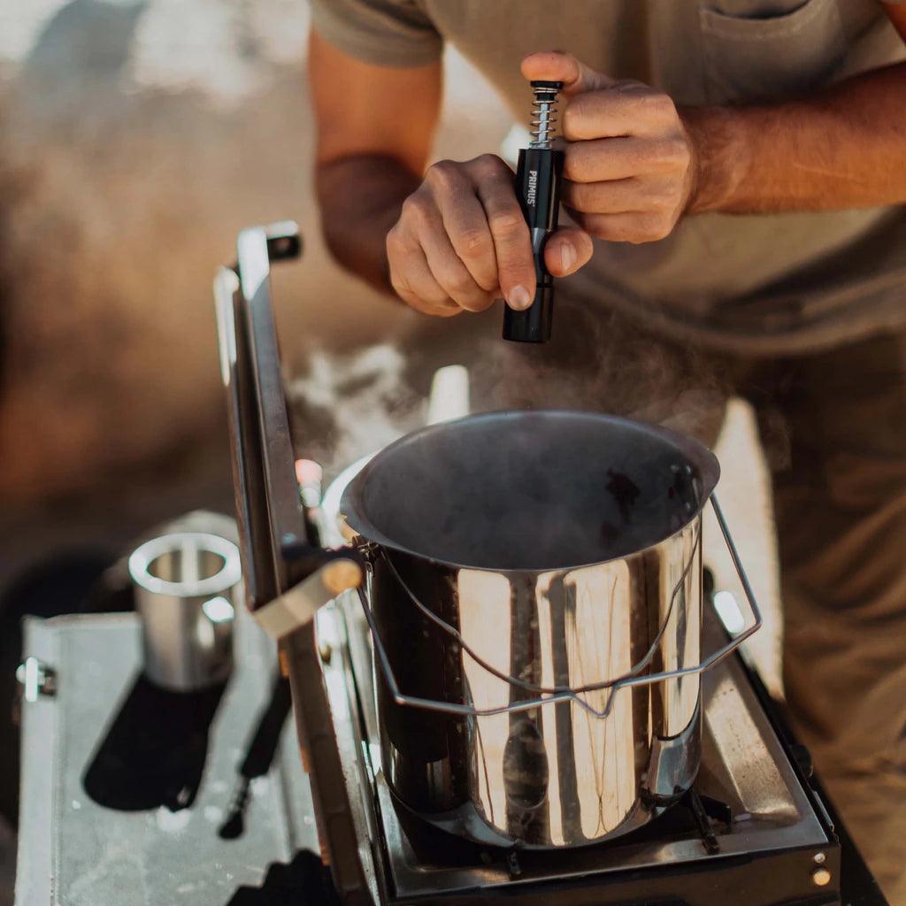 Primus stainless steel campfire pot on outdoor stove emitting steam, person lighting stove