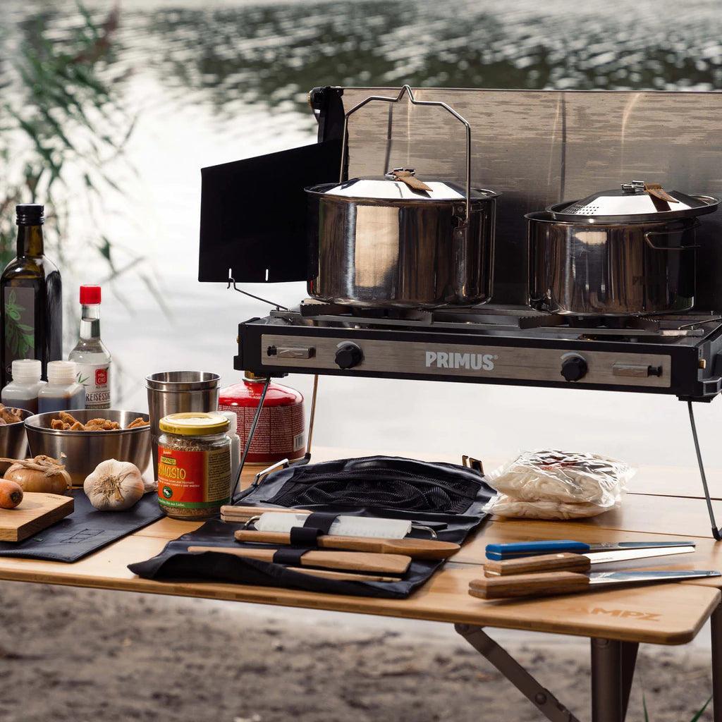 Primus campfire prep set with cooking utensils, pots, and ingredients on outdoor table by lake
