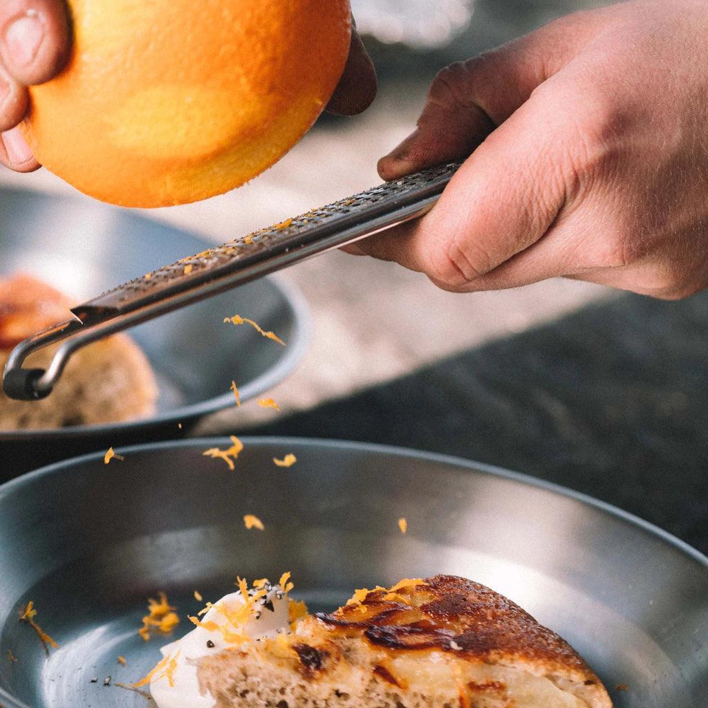 Hand grating orange zest over dessert on stainless steel camping plate outdoors