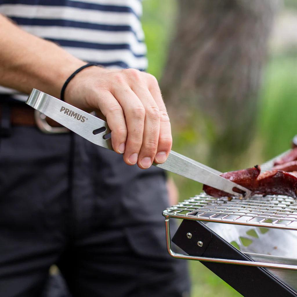 Person using Primus campfire tongs to grill meat outdoors on a metal grate