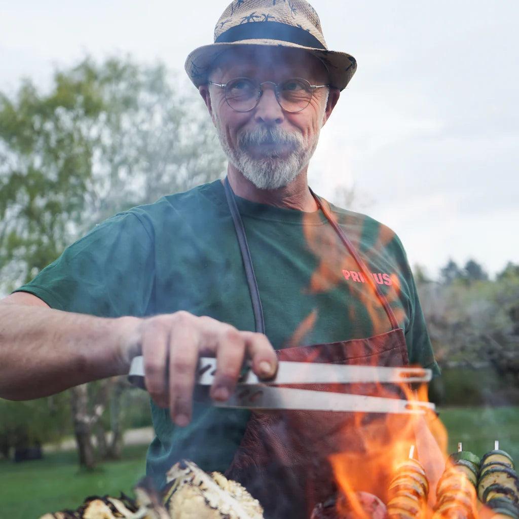 Man grilling outdoors with Primus campfire tongs, wearing a hat and apron, over open flames