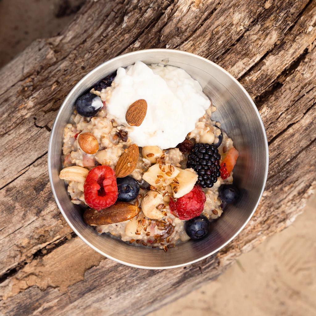 Stainless steel bowl with oatmeal, nuts, and berries on rustic wood outdoors