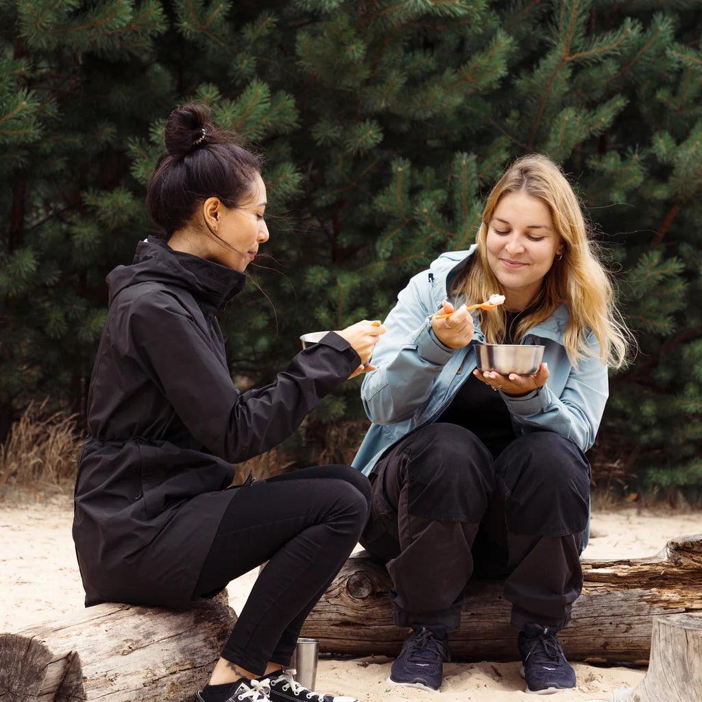 Two women eating from stainless steel bowls outdoors on a log, campsite setting with pine trees.